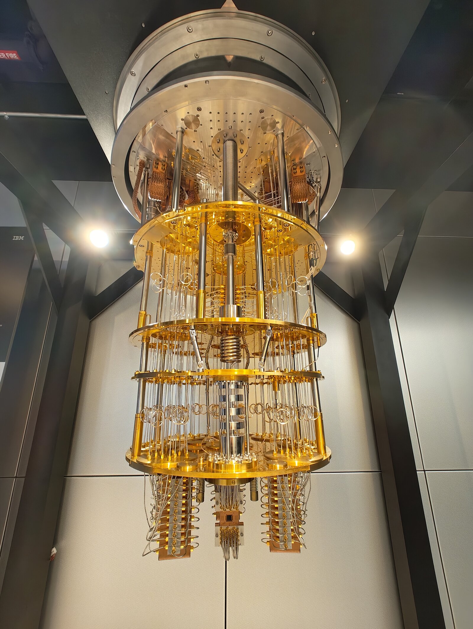 A quantum computer is mostly this: the gold “chandelier” is a dilution refrigerator that cools the chip to near absolute zero, with layers of wiring carrying microwave control pulses down to the qubits at the bottom. The actual processor is a fingernail-sized chip; everything else keeps it cold and calibrated. Photo: OJB Quantum, CC BY 4.0.